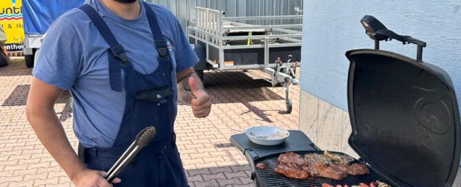 Ein Mann in Blaumann steht am Grill und brät Bratwürste und Steaks mit einem Bratwurstbratgerät.