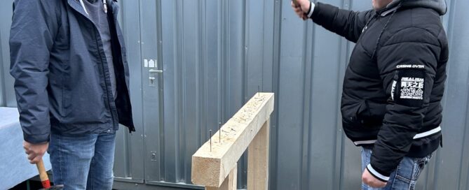 Zwei Mitarbeiter der Firma Günther treten beim Wettnageln gegeneinander an und hauen mit einem Hammer auf Nägel in einen Holzbalken.